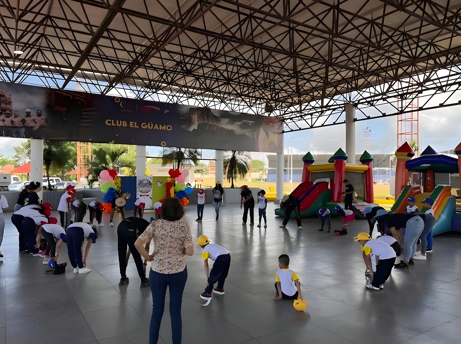 Children in white shirts and yellow caps do guided exercises on a covered court, with bouncy castles behind.