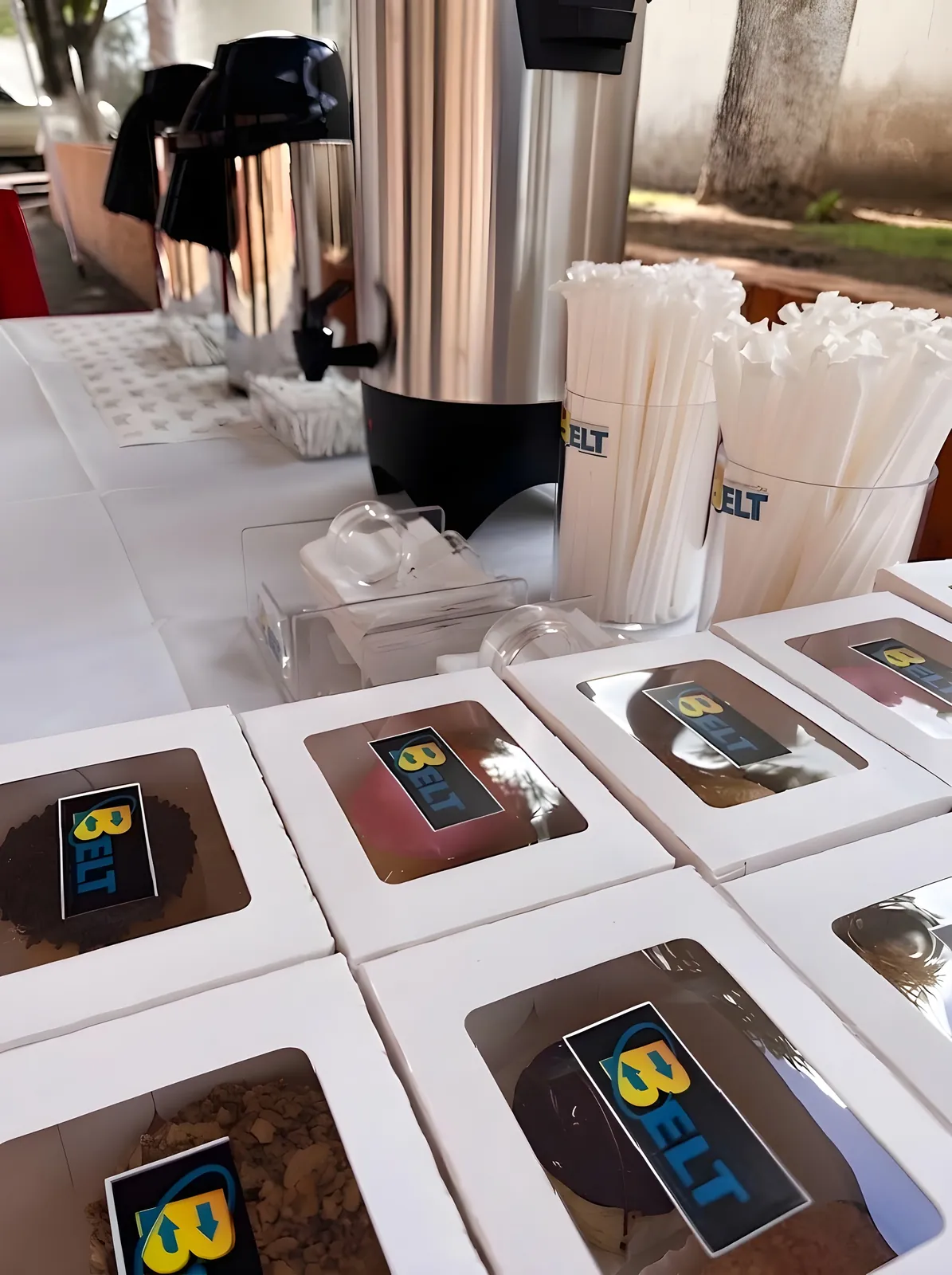 Mesa con cajas de postres, vasos de pitillos con la marca BELT y termos de café.