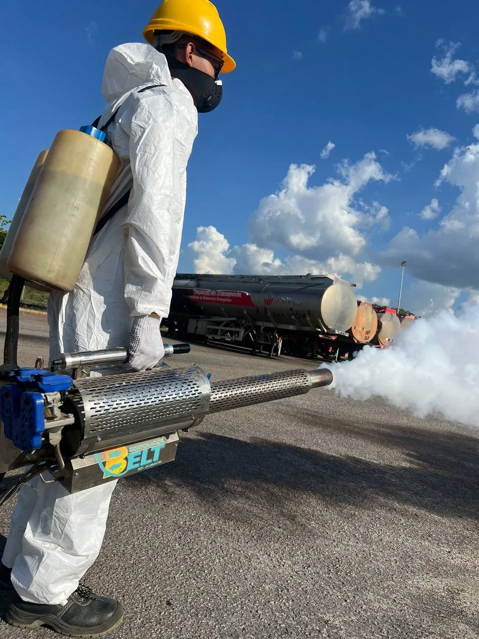 Worker in white coveralls, yellow hard hat and respirator operates a thermal fogger bearing the BELT logo; parked tankers behind.